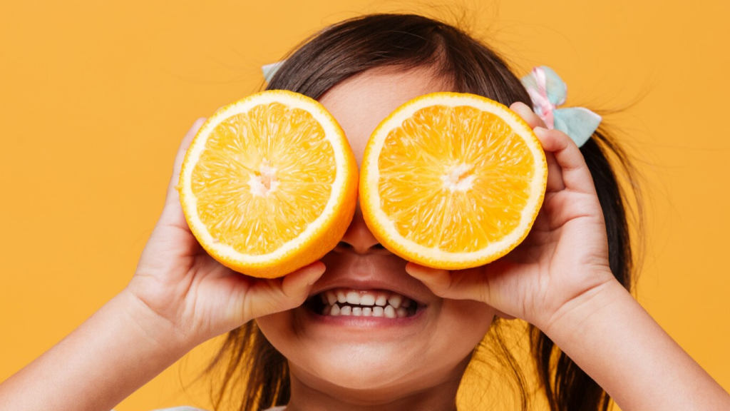 Menina sorrindo e segurando duas metades de laranja na frente do seus olhos, para falar sobre a relação entre a Vitamina D e epilepsia